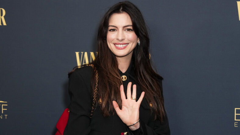 Anne Hathaway in a black jacket, waving at camera
