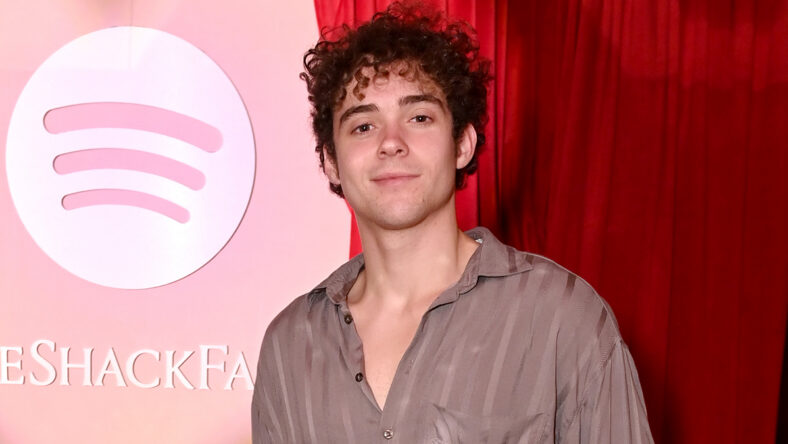 Joshua Bassett poses in front of a pink Spotify backdrop and red curtain while wearing a loose striped button-up shirt.
