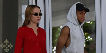 Kylian Mbappé and Ester Expósito, him in a grey sleeveless hoddie and her in a red top, walk outside of a hotel together