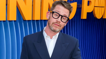 Macaulay Culkin tilts his head to the side while looking at the camera. He's wearing glasses and a suit.