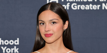 Olivia Rodrigo on a grey background with white writing, wearing red lipstick and a light smile