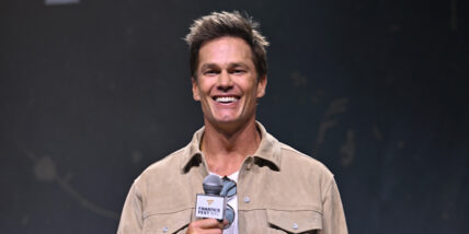 Tom Brady holds a microphone in front of a black background while smiling
