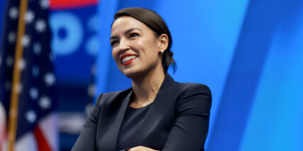 Alexandria Ocasio-Cortez standing in front of a blue background, smiling with the American flag peeking from the left side