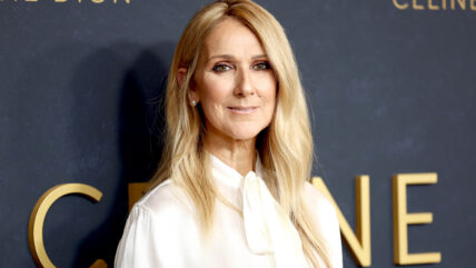 A medium close-up of Celine Dion smiling at the camera with her blonde hair down. She's wearing a white blouse and attending screening of her documentary.