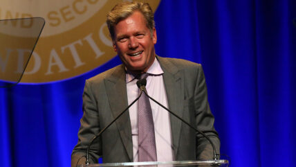 Chris Hansen in a suit stands at a podium with a microphone in front of him.
