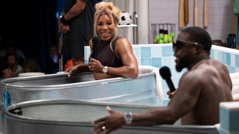 Serena Williams and Kevin Hart sit in metal tubs filled with ice water, holding microphones and smiling, appearing to have a conversation in a studio with sports equipment and blue-tiled walls in the background