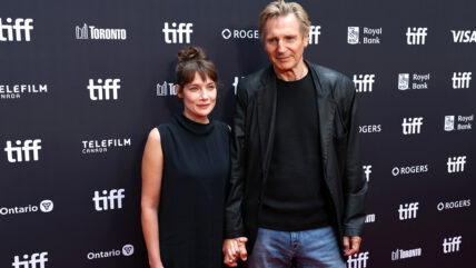 Liam Neeson and Stella Stocker hold hands at Evil Genius premiere at TIFF. Liam in a black blazer and jeans, Stella in a black cowl-necked dress.