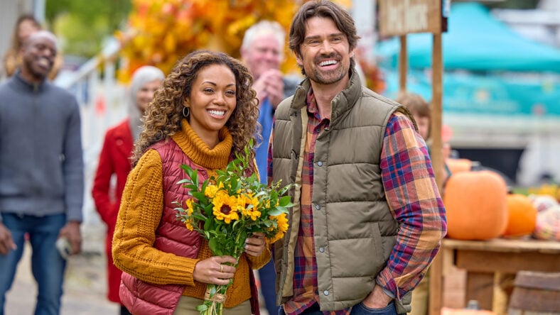 Mishael Morgan and Corey Sevier pose together in a fall-themed setting for a still from Pumpkin Regatta Romance. Mishael is holding a bouquet of yellow flowers.