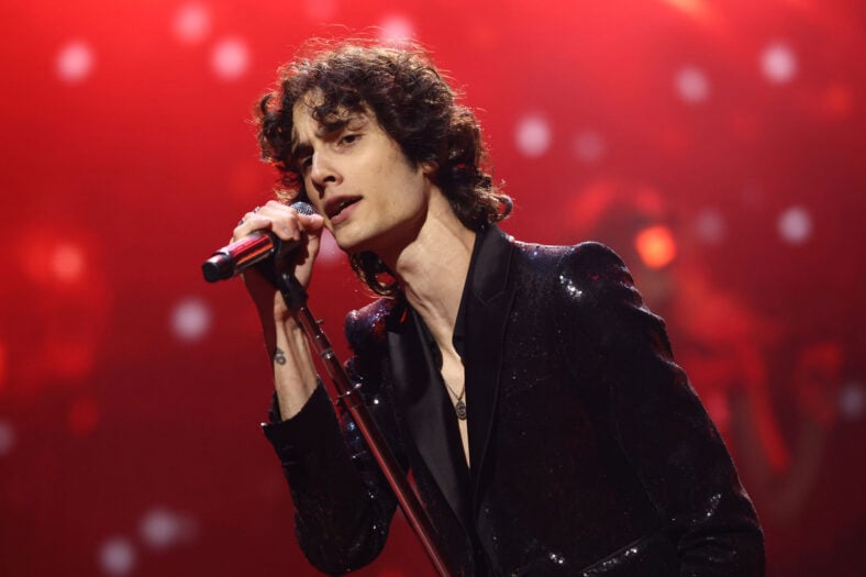 Sombr performing on stage against a red background, holding a microphone