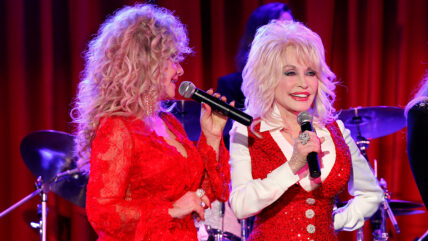 Stella Parton and Dolly Parton perform on stage, both wearing red outfits.