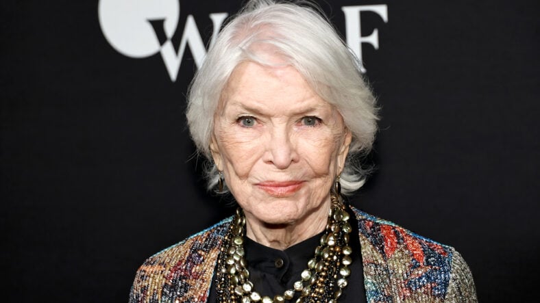 Ellen Burstyn at an event. She has short, white hair and is wearing a multicolored jacket over a black shirt, with layered gold and dark beaded necklaces against a black background.