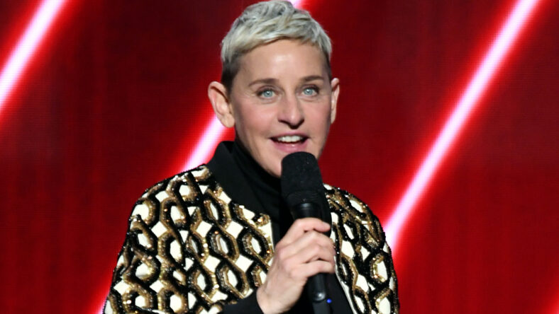 Ellen DeGeneres performs on stage with a microphone in front of a red backdrop.