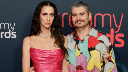 Hila Klein and Ethan Klein pose together on a red carpet in front of a dark backdrop featuring text, with Hila wearing a metallic pink dress and chain necklace, and Ethan wearing a multi-colored patterned shirt.