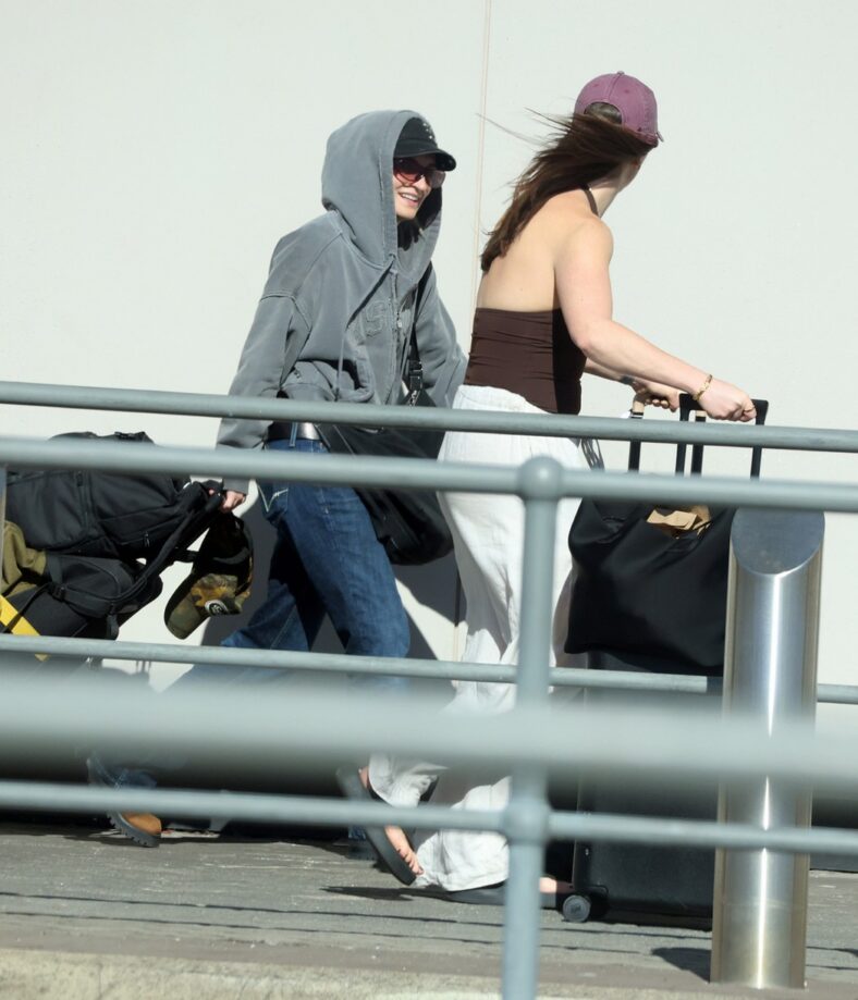 G Flip walks through Perth Airport wearing a gray hooded sweatshirt, sunglasses, and a black cap, smiling alongside a companion pushing a cart filled with luggage.