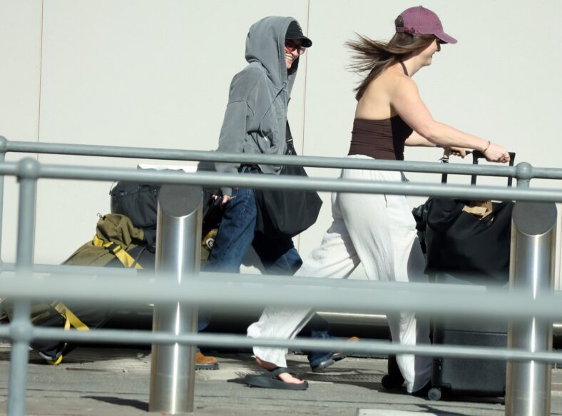 G Flip smiles while walking outside the airport terminal in a gray hoodie and sunglasses, pulling luggage behind a friend who is wearing a maroon baseball cap and white pants.