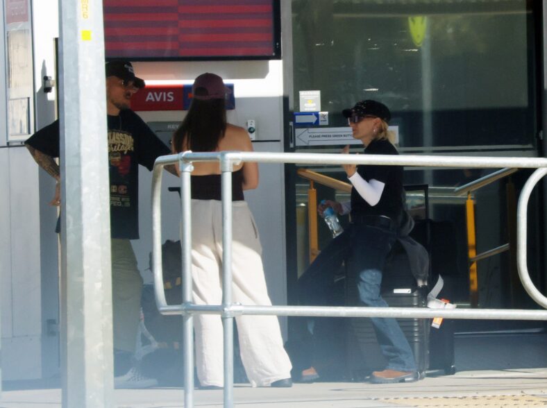 G Flip stands outside a car rental office at the airport, chatting with two companions while wearing a black t-shirt, white undersleeves, jeans, and a black cap.