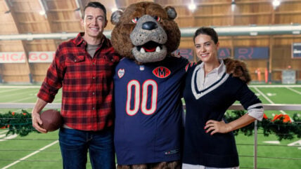 Robert Buckley and Lyndsy Fonseca pose with Staley Da Bear, the Chicago Bears mascot, on an indoor football field for Hallmark's Holiday Touchdown.