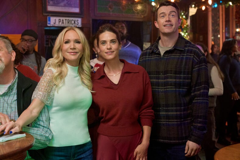 Barbara Alyn Woods, Lyndsy Fonseca, and Robert Buckley stand together inside a crowded, festively decorated pub for a scene in Hallmark's Holiday Touchdown.