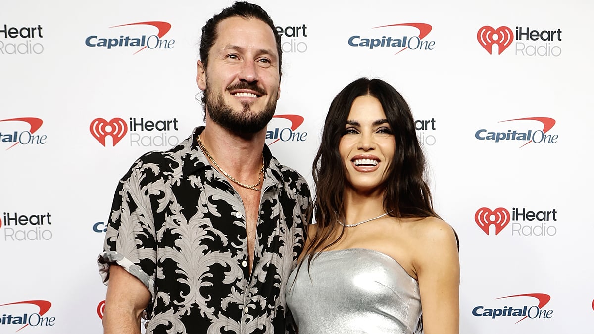 A smiling Val Chmerkovskiy with a slicked-back ponytail and a patterned black-and-grey shirt posing next to Jenna Dewan.