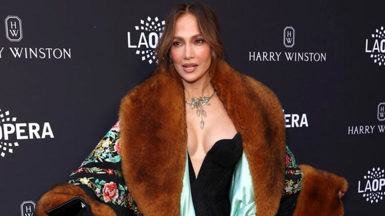 Jennifer Lopez poses on a red carpet wearing a black jumpsuit and a patterned coat with large brown fur trim.