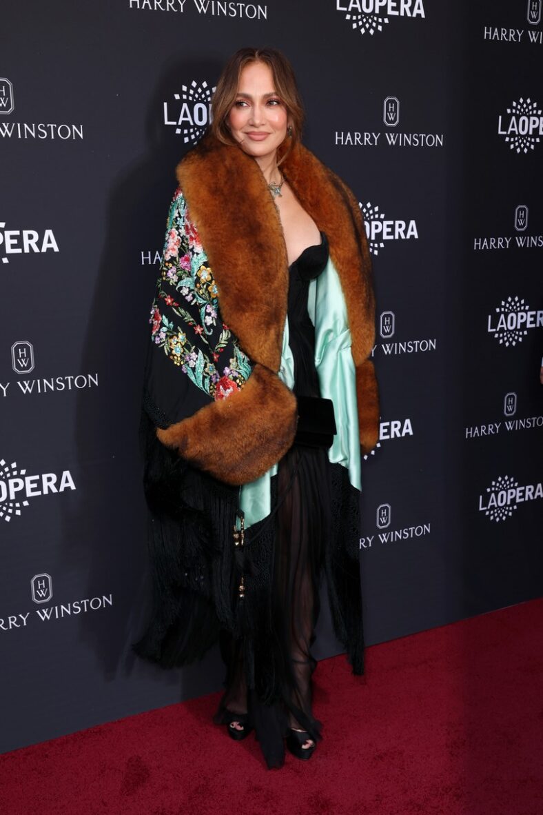 Jennifer Lopez poses on a red carpet wearing a black jumpsuit and a patterned coat with large brown fur trim.