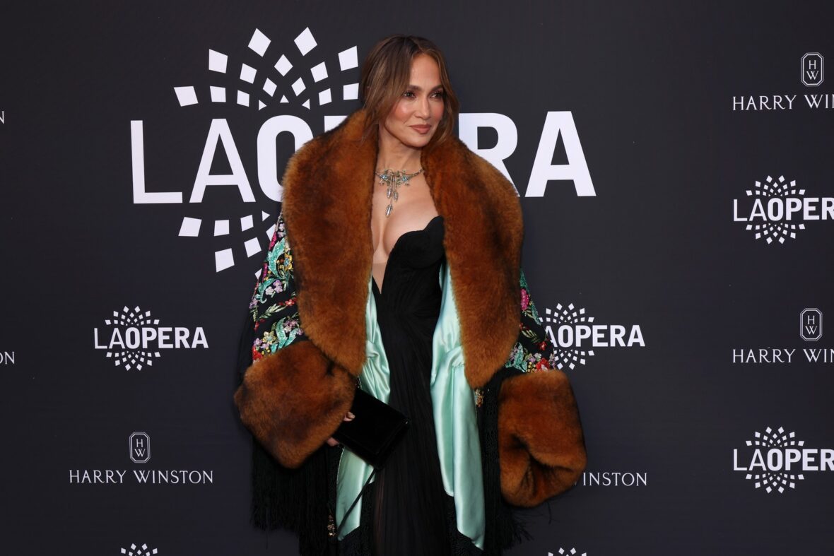 Jennifer Lopez poses on a red carpet wearing a black jumpsuit and a patterned coat with large brown fur trim.