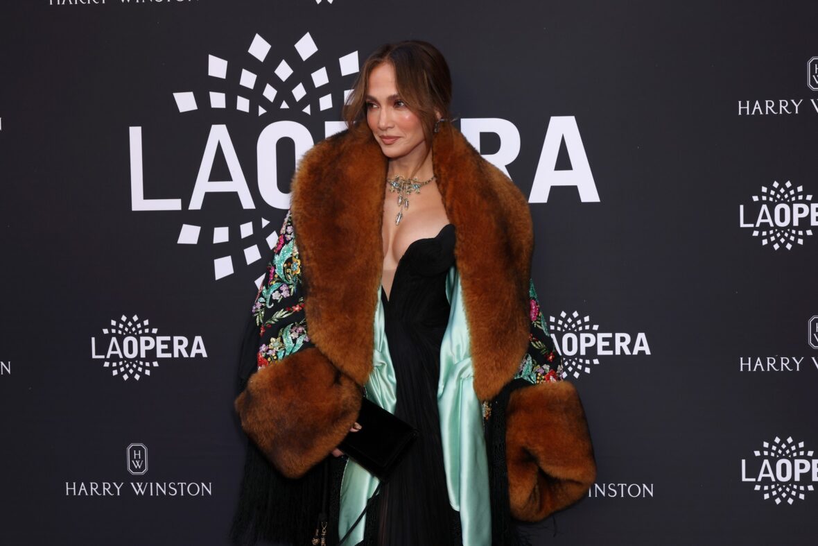 Jennifer Lopez poses on a red carpet wearing a black jumpsuit and a patterned coat with large brown fur trim.