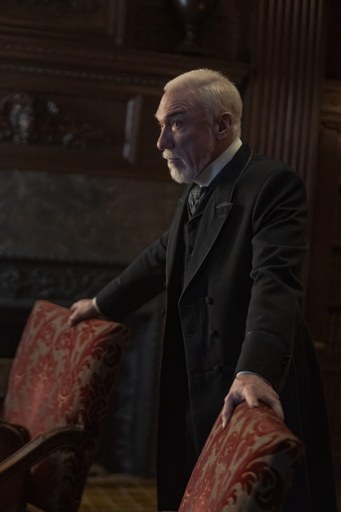 Patrick Page as Richard Clay stands in profile, gripping the back of a red patterned armchair while wearing a dark period coat.
