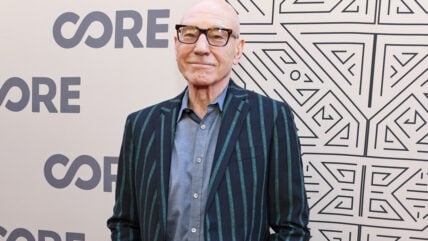 Actor Sir Patrick Stewart smiles while standing in front of a step-and-repeat backdrop featuring the word "CORE" and black-and-white geometric patterns. He wears tortoiseshell-rimmed glasses, a grey button-down shirt, and a dark blazer with vertical teal stripes.