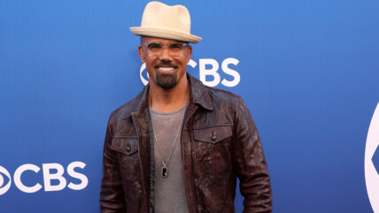 A medium shot of actor Shemar Moore smiling on a blue carpet in front of a CBS backdrop. He's wearing a dark brown leather jacket over a grey t-shirt, a long silver necklace, square-framed eyeglasses, and a tan fedora. He has a goatee and mustache.