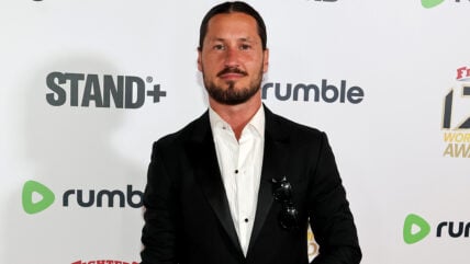 Val Chmerkovskiy smiles while posing on a red carpet in a black suit jacket, an open-collared white dress shirt, and sunglasses tucked into his front pocket.