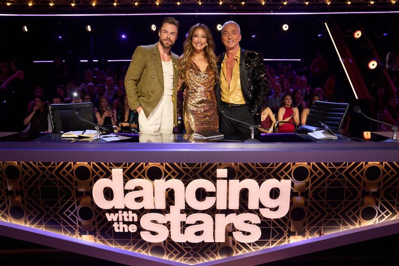 Carrie Ann Inaba, Derek Hough, and Bruno Tonioli pose for a photo behind the judges desk on DWTS