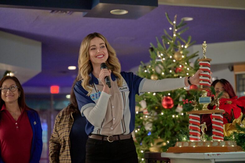 Ashley Newbrough in a blue and beige bowling shirt speaks into a microphone while holding a large multi-tiered holiday bowling trophy next to a Christmas tree.