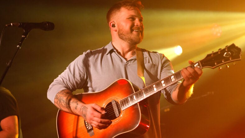 Zach Bryan performing on stage with an acoustic guitar.